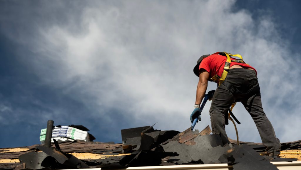 A roofer tears off damaged shingles against a cloudy sky.
