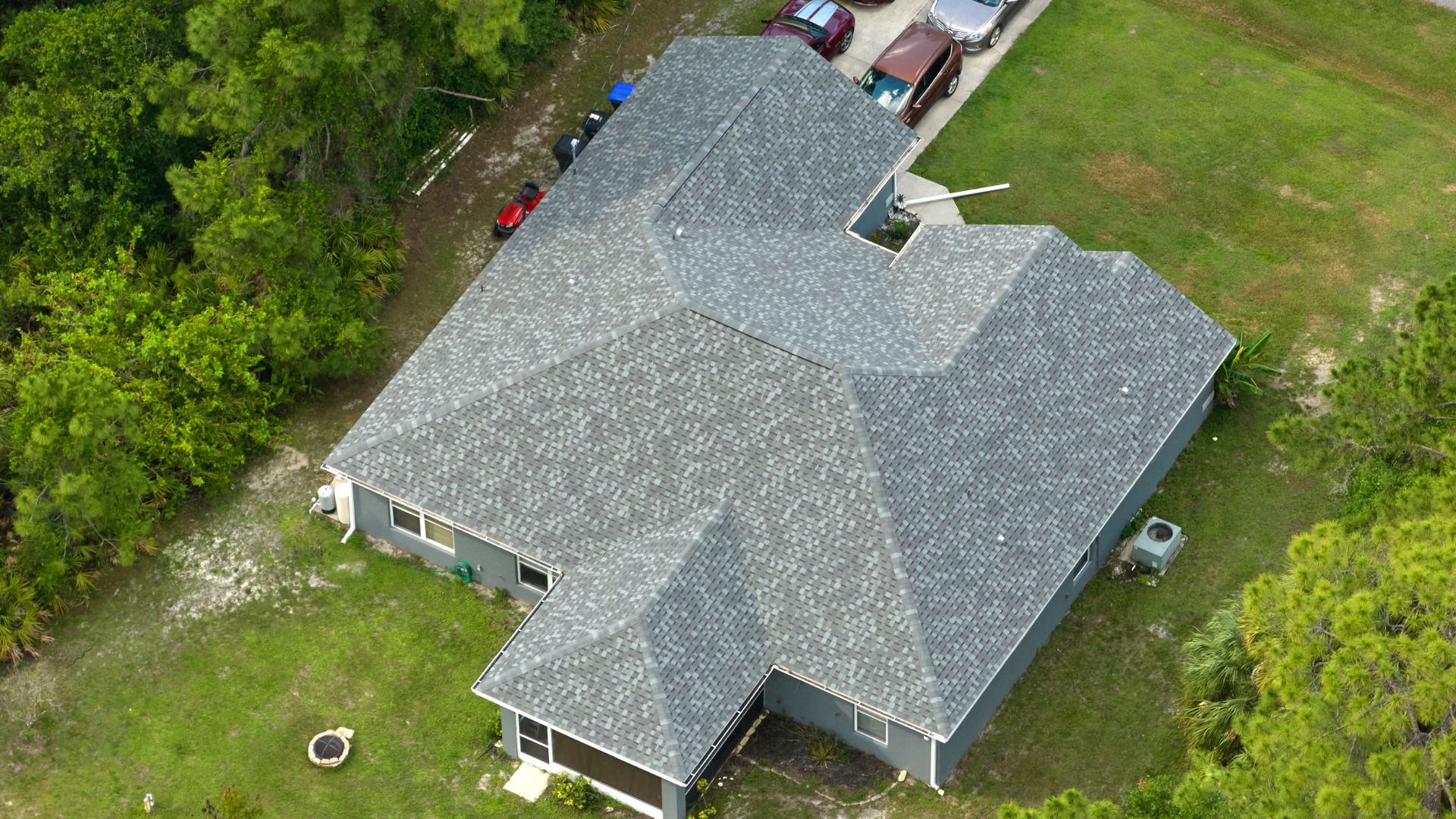 An aerial view of the asphalt shingle roof of a suburban home surrounded by trees and greenery.