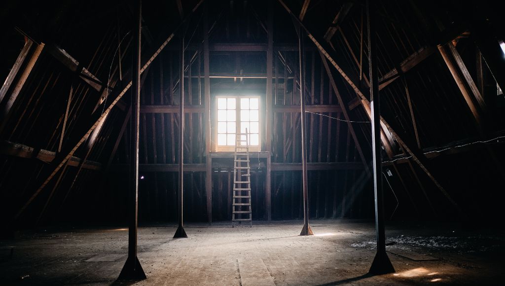 The bare-bones attic of a house with exposed beams and a ladder leading to a window.